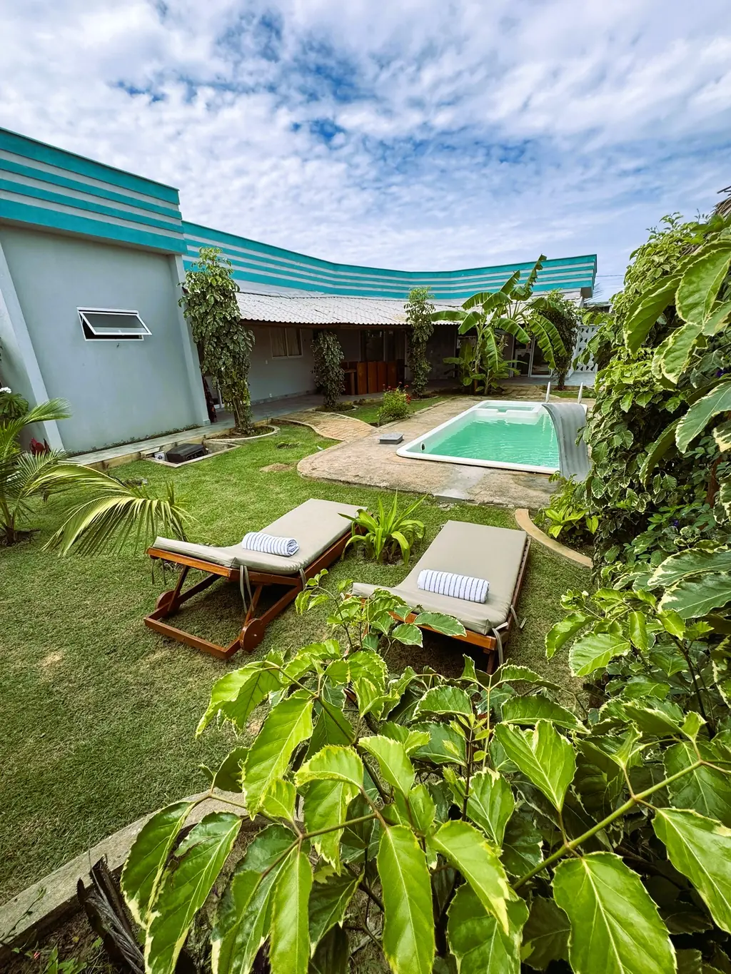 Rectangular pool with two sun loungers on the lawn, tropical foliage in front and the striped parapet behind
